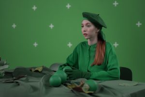 A woman dressed in green, wearing a beret and gloves, sits at a table with green objects.